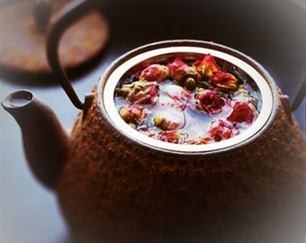 May include: A close-up of a brown teapot filled with water and rose buds. The teapot has a textured surface and a silver rim. The lid is slightly ajar, revealing the contents. The tea is a light pink color.