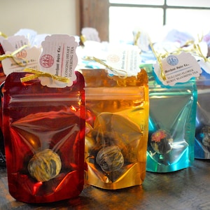 May include: Assortment of colorful, metallic pouches containing loose leaf tea. Each pouch has a tag with the name of the tea, such as "Volcano Flower Ruby Blooming Tea" and "Longing Heart Flowering Tea".
