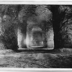 K&ouml;nnte beinhalten: Ein Schwarzwei&szlig;foto eines gro&szlig;en, h&ouml;hlenartigen Raumes mit einer rauen, felsigen Decke und W&auml;nden. Der Raum wird von einer einzigen Lichtquelle in der Ferne beleuchtet, die lange Schatten wirft. Eine Person steht im Vordergrund und wird von der Weite des Raumes &uuml;berragt.