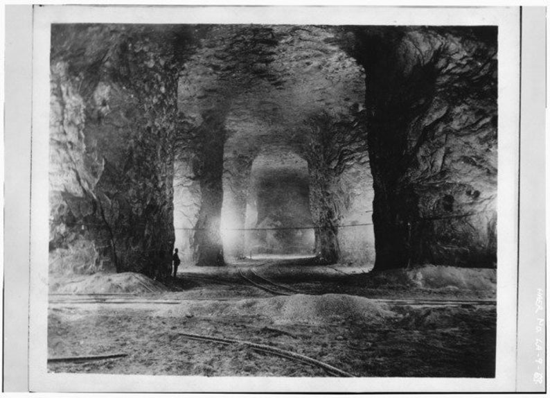 K&ouml;nnte beinhalten: Ein Schwarzwei&szlig;foto eines gro&szlig;en, h&ouml;hlenartigen Raumes mit einer rauen, felsigen Decke und W&auml;nden. Der Raum wird von einer einzigen Lichtquelle in der Ferne beleuchtet, die lange Schatten wirft. Eine Person steht im Vordergrund und wird von der Weite des Raumes &uuml;berragt.