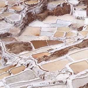 May include: Aerial view of a salt flat with hundreds of individual salt pans, each filled with a shallow layer of water. The pans are arranged in a grid pattern and are surrounded by white salt deposits. The water in the pans is a light brown color, reflecting the sunlight.