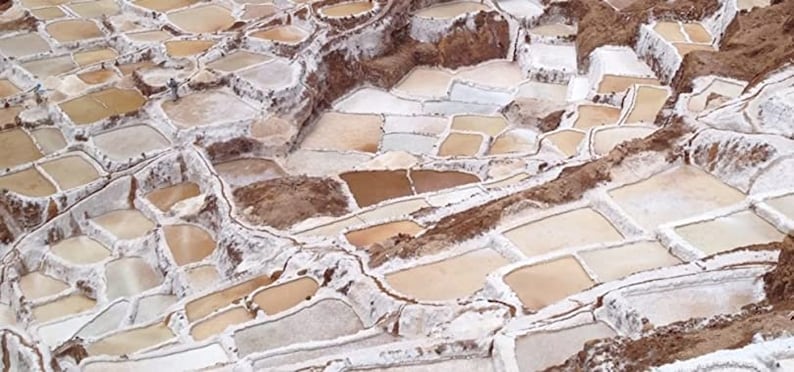 May include: Aerial view of a salt flat with hundreds of individual salt pans, each filled with a shallow layer of water. The pans are arranged in a grid pattern and are surrounded by white salt deposits. The water in the pans is a light brown color, reflecting the sunlight.