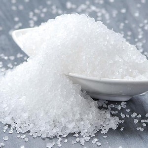 May include: A close-up of a white, fine-grained salt, piled high in a white ceramic bowl. The salt is spilling out of the bowl onto a gray surface.