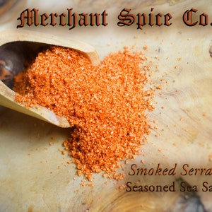 May include: A wooden scoop filled with orange-red smoked serrano sea salt, spilling onto a wooden surface. The text "Merchant Spice Co." is visible at the top of the image. The text "Smoked Serrano Sea Salt" and "Seasoned Sea Salt Collection" is visible at the bottom of the image.