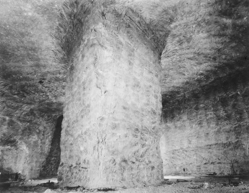 K&ouml;nnte beinhalten: Ein Schwarzwei&szlig;foto eines gro&szlig;en, grob behauenen Steinpfeilers in einem h&ouml;hlenartigen Raum. Der Pfeiler ist hoch und breit, und die umliegenden W&auml;nde sind rau und uneben.