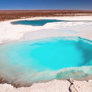 May include: A bright blue lagoon surrounded by white salt flats. The water is crystal clear and the sky is a deep blue.