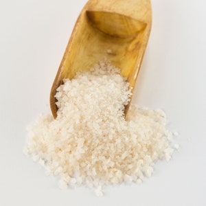 May include: A wooden scoop filled with coarse white sea salt, spilling out onto a white surface.