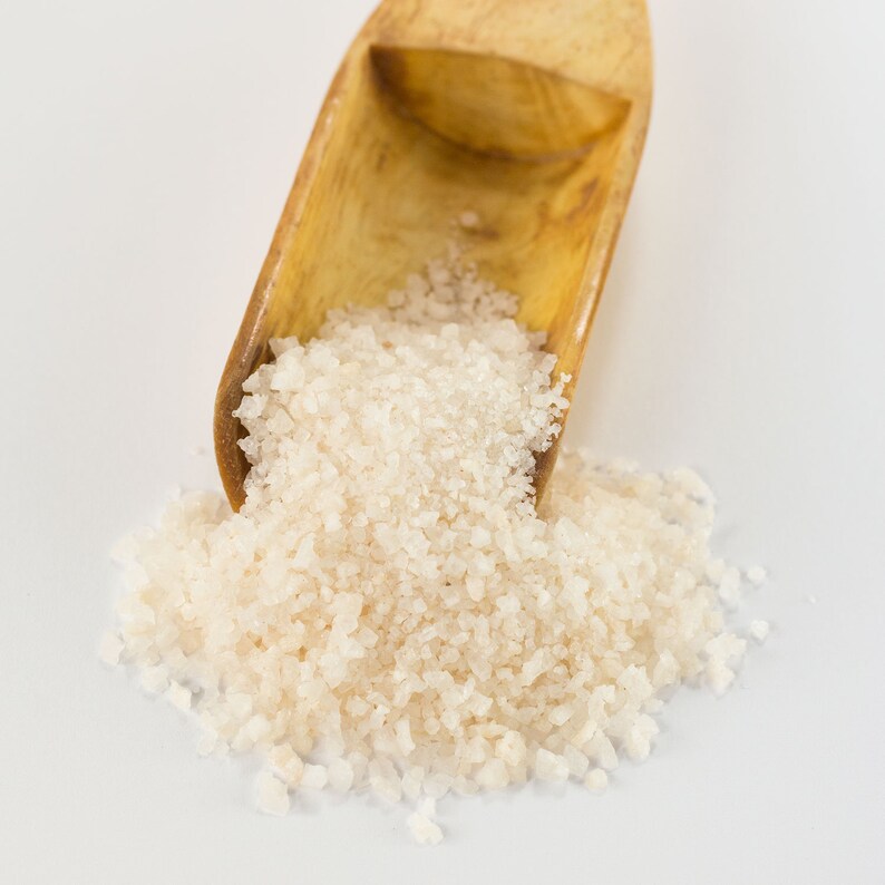 May include: A wooden scoop filled with coarse white sea salt, spilling out onto a white surface.