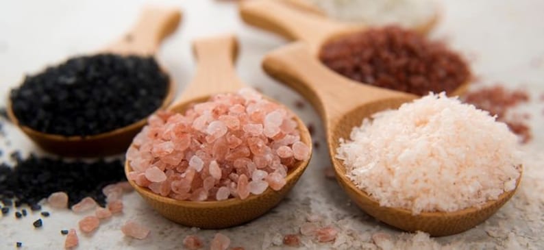 May include: Five wooden spoons filled with different types of salt: black salt, pink Himalayan salt, red salt, and two types of white salt.