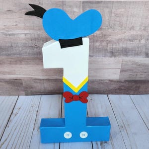 May include: A blue and white number one shaped like Donald Duck. The number has a black hat, a red bow tie, and two white buttons.