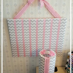 May include: A gray and pink chevron patterned headband organizer with a pink ribbon and bow. The organizer is mounted on a wall and has a matching pink and gray chevron patterned headband holder on a gray base.