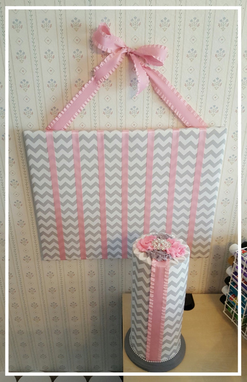 May include: A gray and pink chevron patterned headband organizer with a pink ribbon and bow. The organizer is mounted on a wall and has a matching pink and gray chevron patterned headband holder on a gray base.