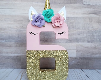 Peut inclure: Numéro six en paillettes roses avec une moitié inférieure en paillettes dorées. Le numéro a une corne de licorne, des oreilles et une couronne de fleurs avec des fleurs roses, violettes et turquoise.
