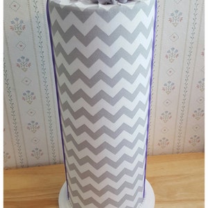 May include: A tall, cylindrical decorative item with a white and gray chevron pattern. It has a white base with a pearl trim and is topped with purple and white fabric flowers. A purple ribbon accents the sides.