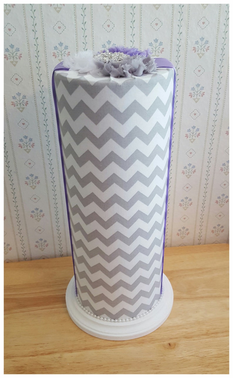 May include: A tall, cylindrical decorative item with a white and gray chevron pattern. It has a white base with a pearl trim and is topped with purple and white fabric flowers. A purple ribbon accents the sides.