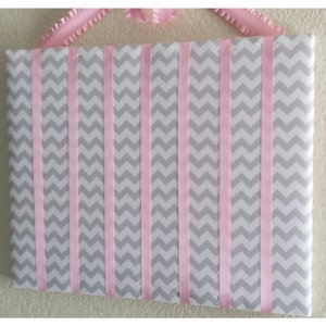 May include: A gray and pink chevron patterned fabric board with pink ribbon strips sewn across the surface. The board is designed for hanging items.
