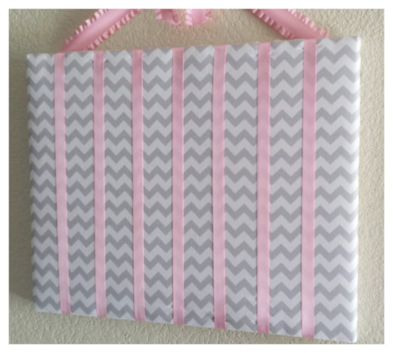 May include: A gray and pink chevron patterned fabric board with pink ribbon strips sewn across the surface. The board is designed for hanging items.