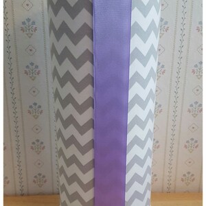 May include: A tall, cylindrical decorative vase with a white base and a gray and white chevron pattern. A wide lavender ribbon is centered on the front. The top is adorned with lavender fabric flowers. The base is decorated with a string of pearls.