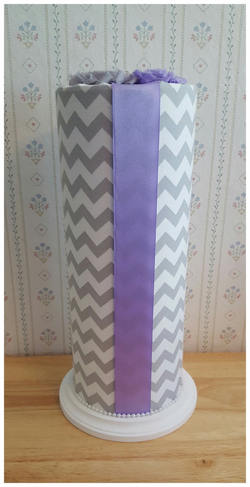 May include: A tall, cylindrical decorative vase with a white base and a gray and white chevron pattern. A wide lavender ribbon is centered on the front. The top is adorned with lavender fabric flowers. The base is decorated with a string of pearls.