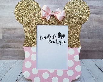 May include: A pink and gold glitter picture frame with a white mat and polka dots. The frame is shaped like Mickey Mouse ears and has a pink bow on top. The text "Kinsley's Bowtique" is printed on the frame.