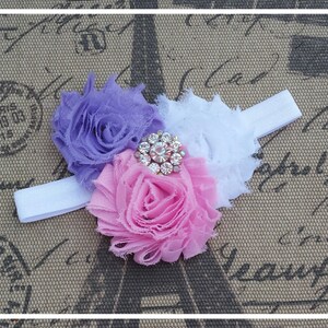 May include: A white elastic headband with three fabric flowers in shades of purple, pink, and white. A sparkly rhinestone embellishment is in the center of the flowers.