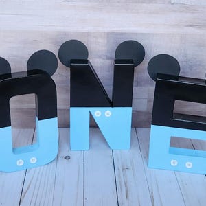 Peut inclure: Lettres en bois noires et bleu clair &eacute;pellant "ONE" avec des oreilles de Mickey Mouse sur chaque lettre. Les lettres sont d&eacute;cor&eacute;es de points blancs.