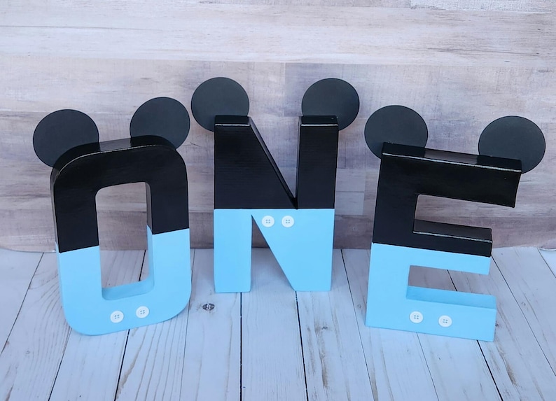 Peut inclure: Lettres en bois noires et bleu clair &eacute;pellant "ONE" avec des oreilles de Mickey Mouse sur chaque lettre. Les lettres sont d&eacute;cor&eacute;es de points blancs.