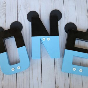 Peut inclure: Trois lettres en bois bleu et noir &eacute;pellant "ONE" avec des oreilles de Mickey Mouse noires et des boutons blancs. Les lettres sont sur un fond en bois blanc.