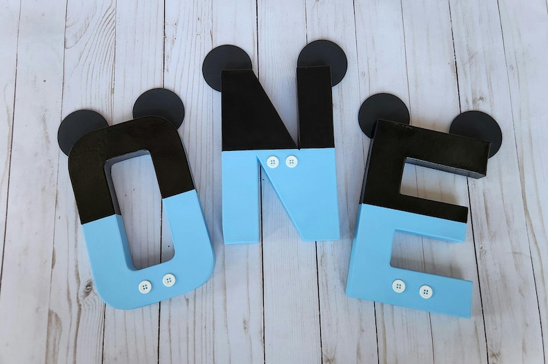Peut inclure: Trois lettres en bois bleu et noir &eacute;pellant "ONE" avec des oreilles de Mickey Mouse noires et des boutons blancs. Les lettres sont sur un fond en bois blanc.