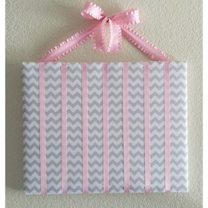 May include: A gray and pink chevron patterned board with pink ribbon strips attached to the top and bottom. The board is hung on a wall with a pink bow.