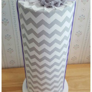 May include: A decorative cylinder with a gray and white chevron pattern. The top is adorned with a cluster of fabric flowers in white, gray, and purple, accented by a silver rhinestone brooch. The base is white with a beaded trim.