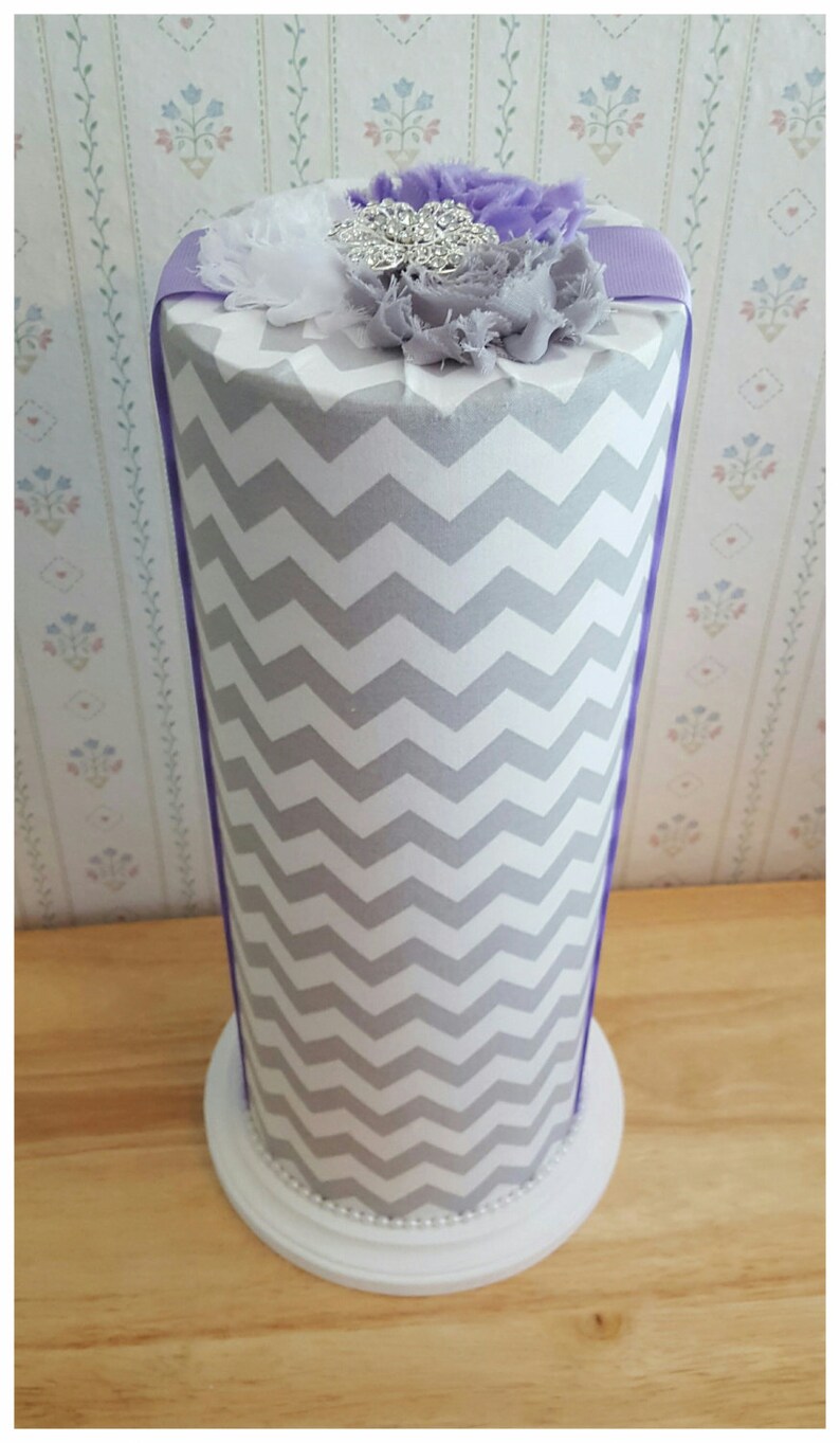 May include: A decorative cylinder with a gray and white chevron pattern. The top is adorned with a cluster of fabric flowers in white, gray, and purple, accented by a silver rhinestone brooch. The base is white with a beaded trim.