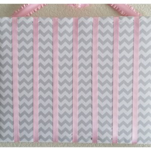 May include: A gray and pink chevron patterned board with pink ribbon strips attached vertically. The board is used for hanging items like headbands or hair clips.