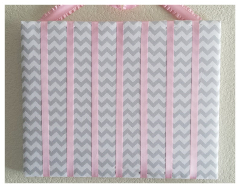 May include: A gray and pink chevron patterned board with pink ribbon strips attached vertically. The board is used for hanging items like headbands or hair clips.