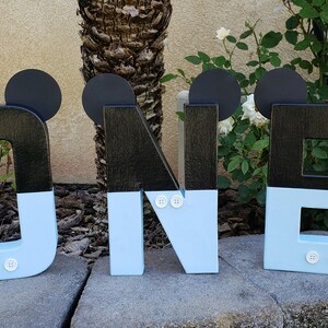 Peut inclure: Lettres en bois noires et bleu clair orthographiant "ONE" avec des oreilles de Mickey Mouse et des boutons blancs. Les lettres sont pos&eacute;es sur une surface en pierre.