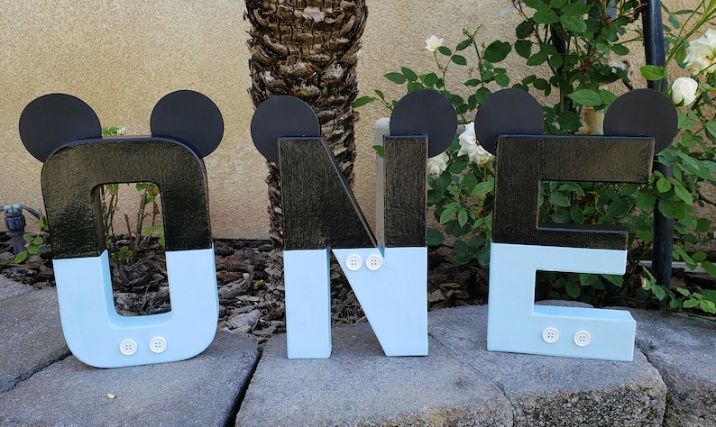 Peut inclure: Lettres en bois noires et bleu clair orthographiant "ONE" avec des oreilles de Mickey Mouse et des boutons blancs. Les lettres sont pos&eacute;es sur une surface en pierre.