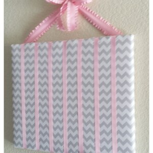 May include: A gray and pink chevron patterned fabric board with pink ribbon strips attached to the top and bottom. The board is designed to hold hair accessories.