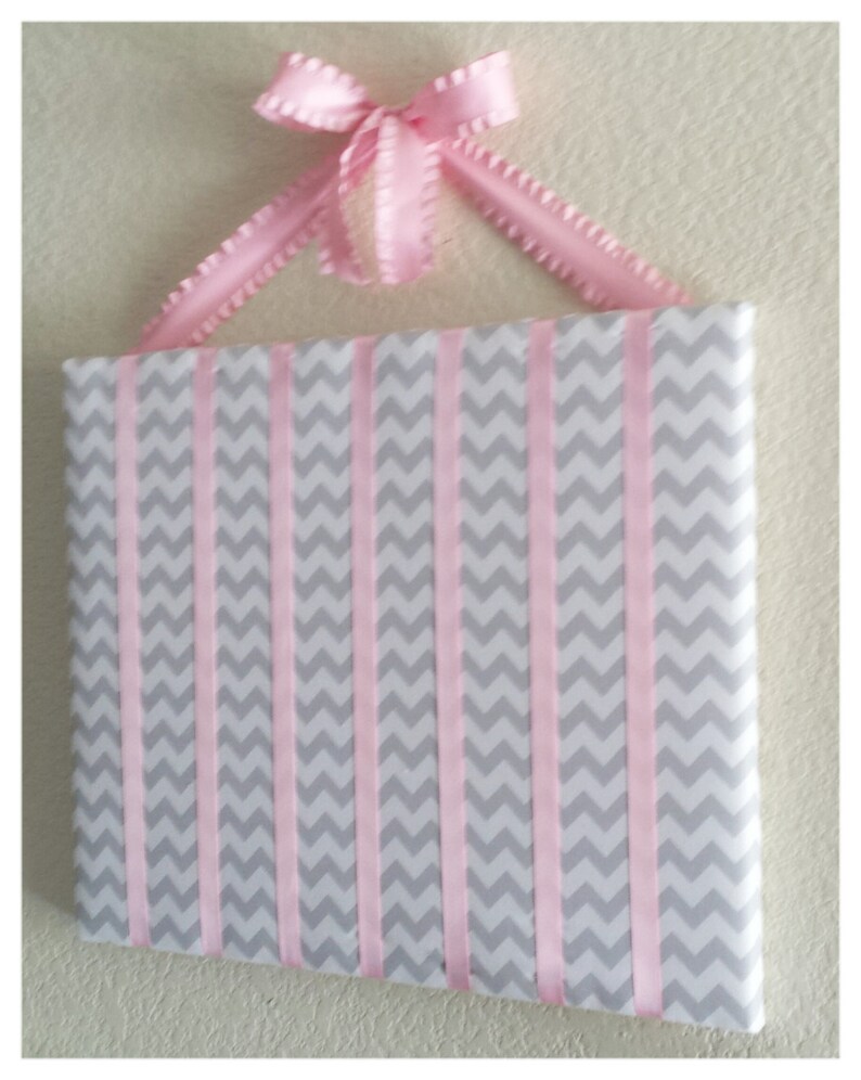May include: A gray and pink chevron patterned fabric board with pink ribbon strips attached to the top and bottom. The board is designed to hold hair accessories.