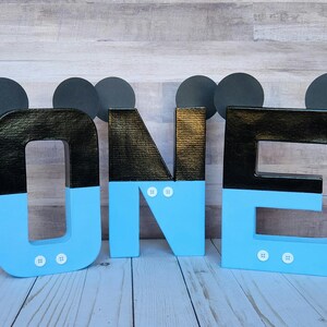 Peut inclure: Lettres en carton noir et bleu &eacute;pellant "ONE" avec des oreilles de Mickey Mouse sur chaque lettre. Les lettres sont d&eacute;cor&eacute;es de boutons blancs.