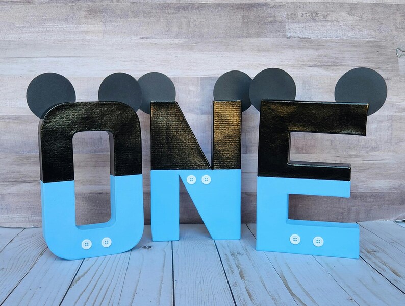Peut inclure: Lettres en carton noir et bleu &eacute;pellant "ONE" avec des oreilles de Mickey Mouse sur chaque lettre. Les lettres sont d&eacute;cor&eacute;es de boutons blancs.