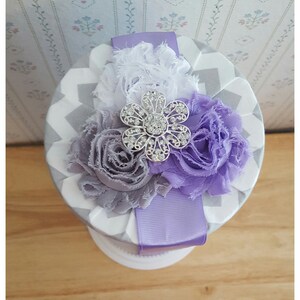 May include: A round, decorative box with a gray and white chevron pattern. The top is adorned with fabric flowers in shades of white, gray, and purple, and a silver flower-shaped embellishment. A lavender ribbon wraps around the box.