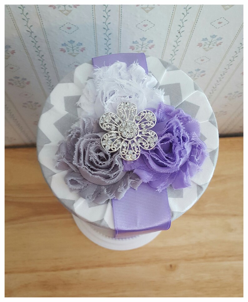 May include: A round, decorative box with a gray and white chevron pattern. The top is adorned with fabric flowers in shades of white, gray, and purple, and a silver flower-shaped embellishment. A lavender ribbon wraps around the box.
