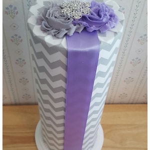 May include: A decorative cylinder with a gray and white chevron pattern, topped with fabric flowers in white, gray, and purple. A silver rhinestone embellishment sits in the center. A wide purple ribbon is affixed to the front.