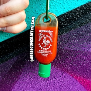 May include: A small, clear plastic bottle filled with orange Sriracha sauce, featuring a green flip-top cap and a green carabiner attached to the top. The label displays the text SRIRACHA2GO, a rooster logo, and the text keep it spicy 1 FL OZ.