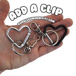 May include: Two heart-shaped keychains, one silver and one black, with a silver cat charm attached to the silver keychain. The text "ADD A CLIP BUBBLEGUMGRAFFITI.COM" is visible in the background.