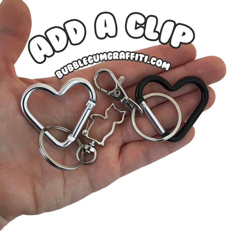 May include: Two heart-shaped keychains, one silver and one black, with a silver cat charm attached to the silver keychain. The text "ADD A CLIP BUBBLEGUMGRAFFITI.COM" is visible in the background.