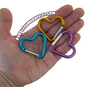 May include: Three heart-shaped metal carabiners in blue, gold, and purple. The carabiners are in the palm of a hand.