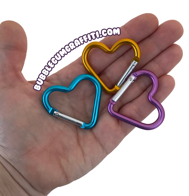 May include: Three heart-shaped metal carabiners in blue, gold, and purple. The carabiners are in the palm of a hand.