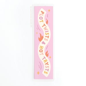 May include: A pink bookmark with the text "PLOT TWISTS & HOT TRYSTS" in a wavy banner design. The bookmark features orange flames and yellow stars. The text and design are in a retro style.