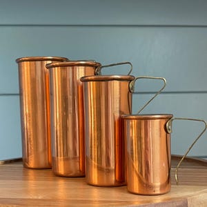 May include: Four copper measuring cups of varying sizes, arranged from largest to smallest. Each cup has a handle and a shiny, reflective surface. The cups are displayed on a wooden surface against a blue background.